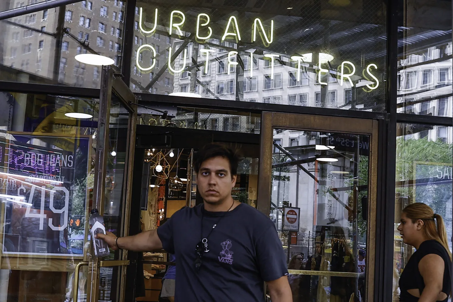 A frowning man holds the door open to an Urban Outfitters.